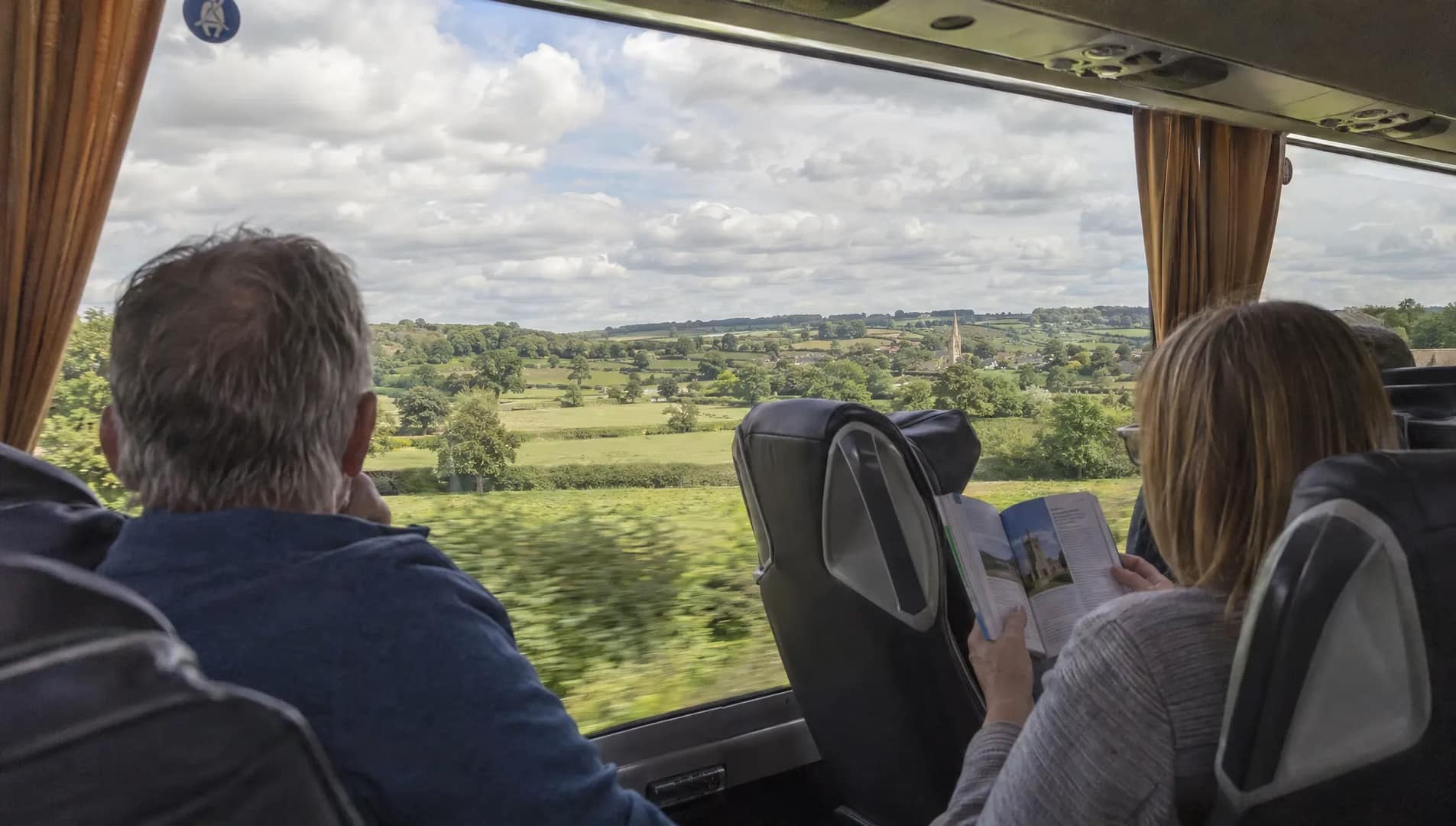 a-passenger-reads-a-guidebook-as-fields-and-hedger-1792685552919 A passenger reads a guidebook as fields and hedgerows pass outside the coach window.