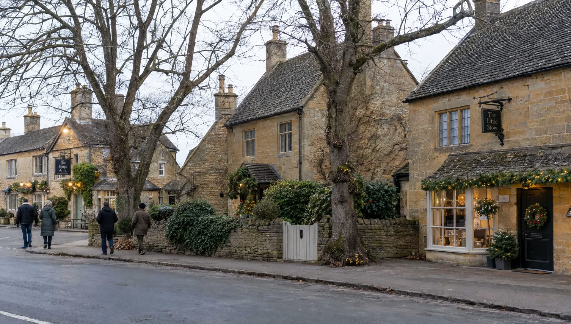 Visitors in warm coats walk along a quiet Cotswolds street lined with stone cottages and bare winter trees on Christmas Day.